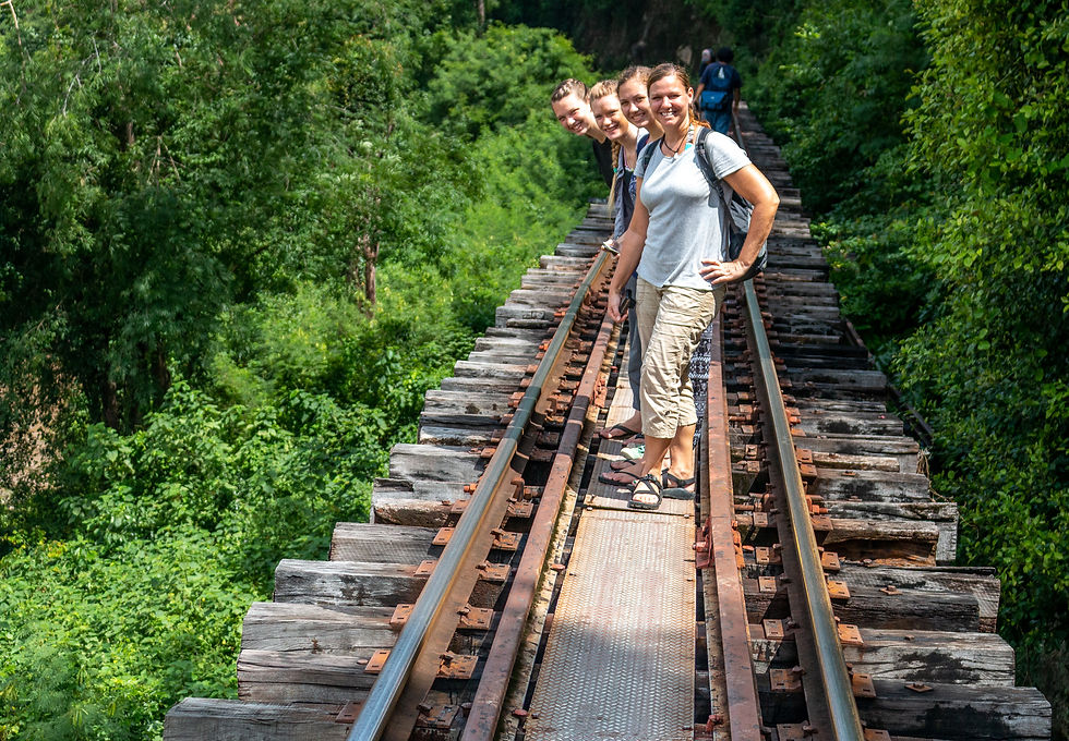 Group of four smiling people stand on a wooden railway track amidst lush greenery, enjoying a sunny day.
