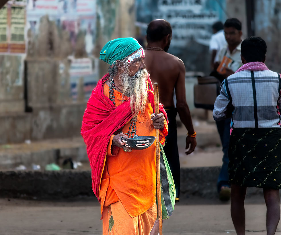 Elderly man with a long beard and colorful robe holds a bowl and staff on a busy street. Posters on wall in the background.