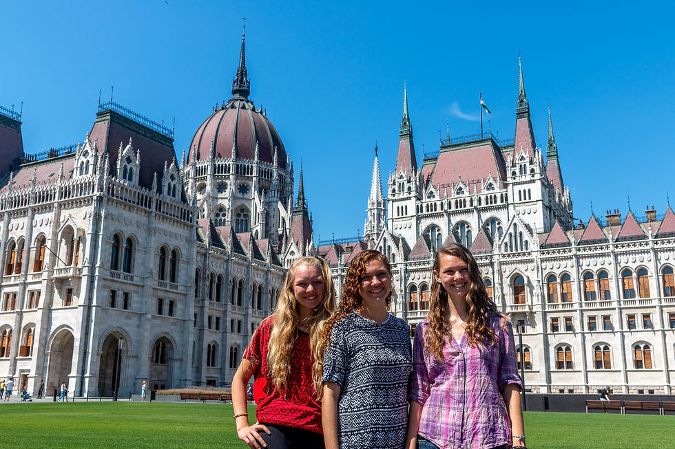 Three smiling women stand on grass in front of a grand, ornate building with spires and a dome under a clear blue sky.