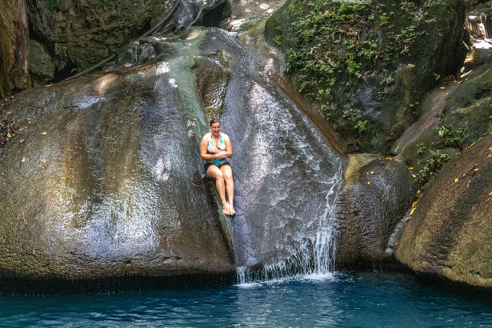 Woman in swimsuit slides down a wet rock into a blue pool, surrounded by lush greenery and sunlight filtering through trees, appearing joyful.