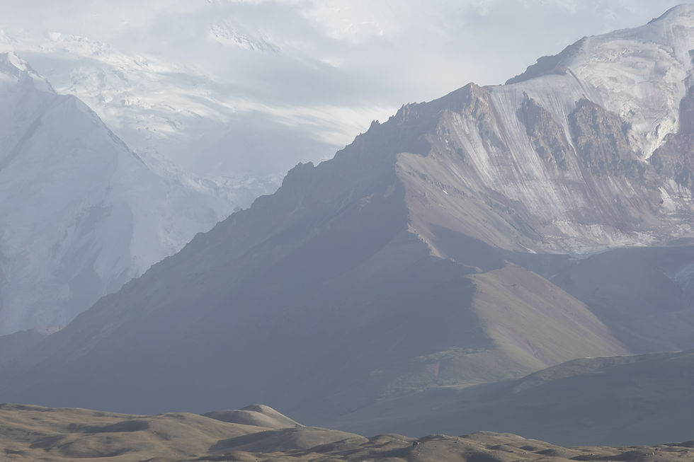Snow-capped mountains under a cloudy sky, with layered rock formations. The foreground features rolling hills in muted earth tones.