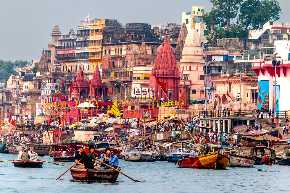 Boats with people on the Ganges River, Varanasi, with colorful buildings and temples in the background. Vibrant flags and busy atmosphere.