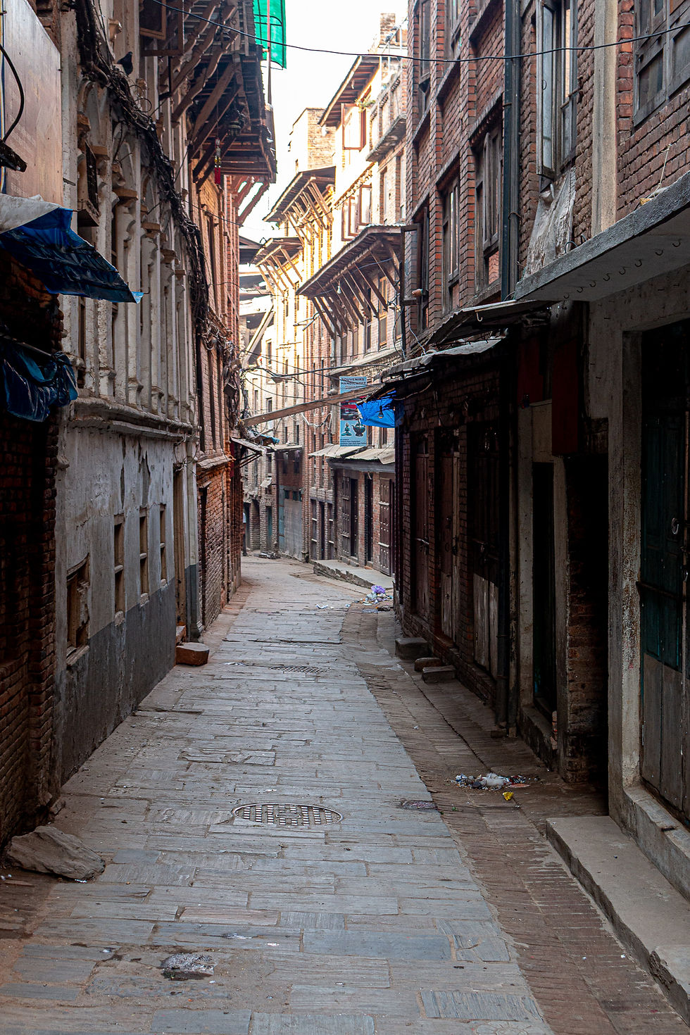 Narrow alley with old brick buildings, sunlight casting shadows, blue tarps, and scattered litter. Quiet, rustic urban scene.