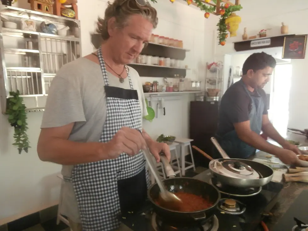 Two men in aprons cook in a kitchen. One stirs sauce, the other prepares ingredients. Shelves with spices and a sign are visible.