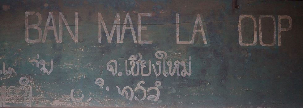 Faded sign with "BAN MAE LA OOP" and Thai text on weathered green wood background, evoking a rustic, historic vibe.