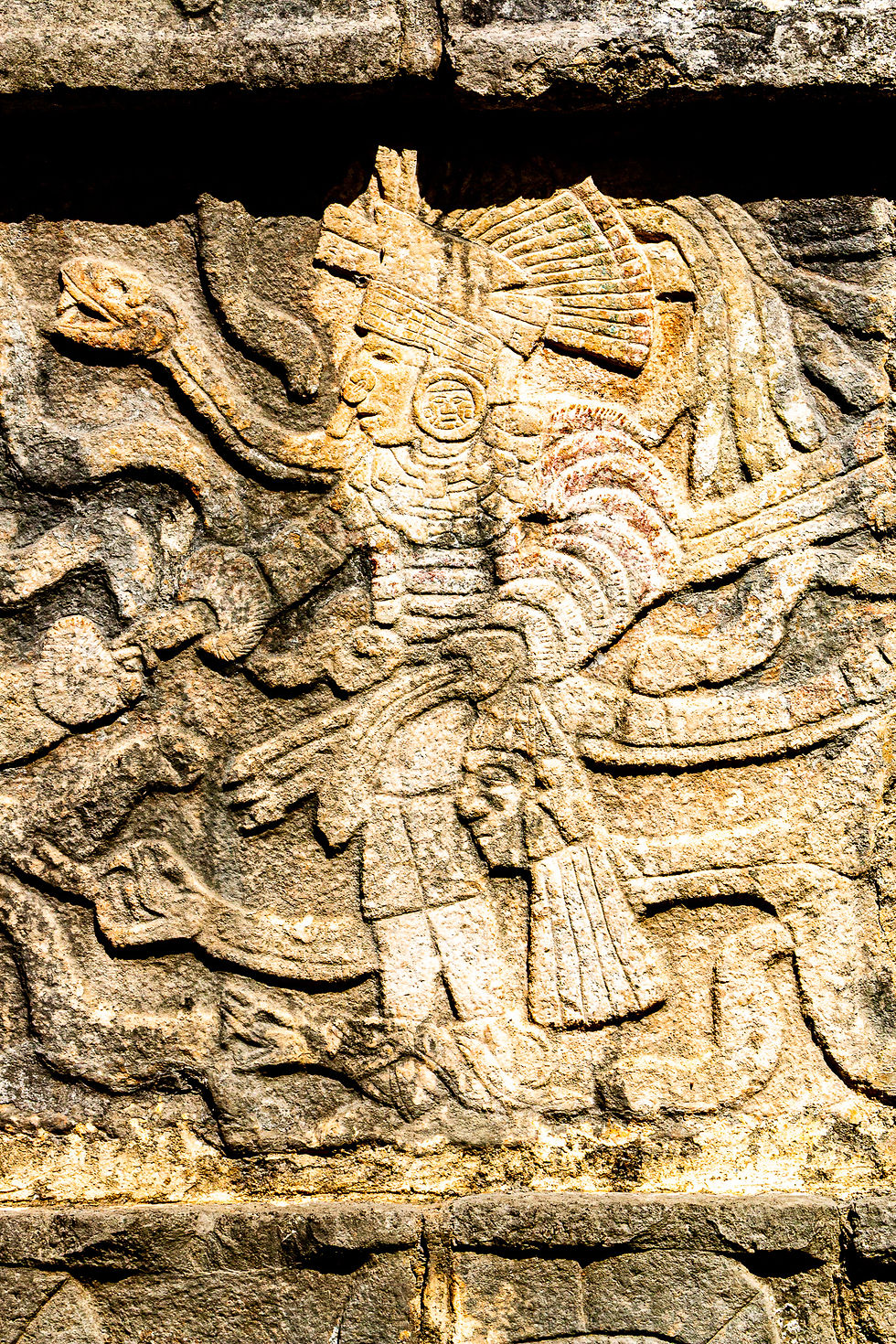 Ancient stone carving of a warrior with a feathered headdress, holding a snake, carved into a weathered stone wall, with intricate details.