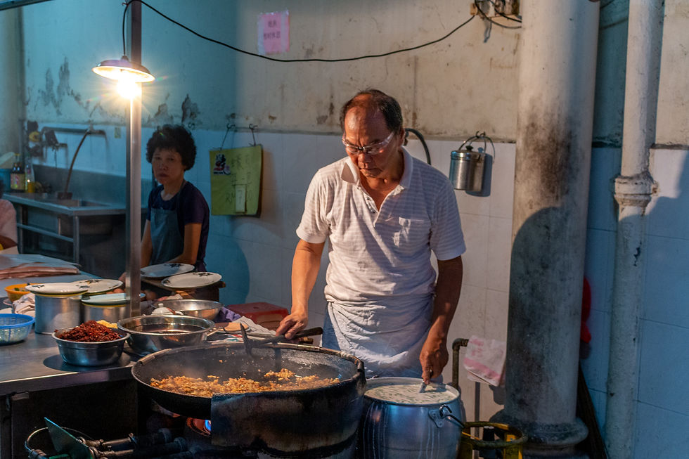 Man in white shirt cooking at a street kitchen under bright light. Woman in apron watches. Pots and utensils on metal counter. Dim setting.
