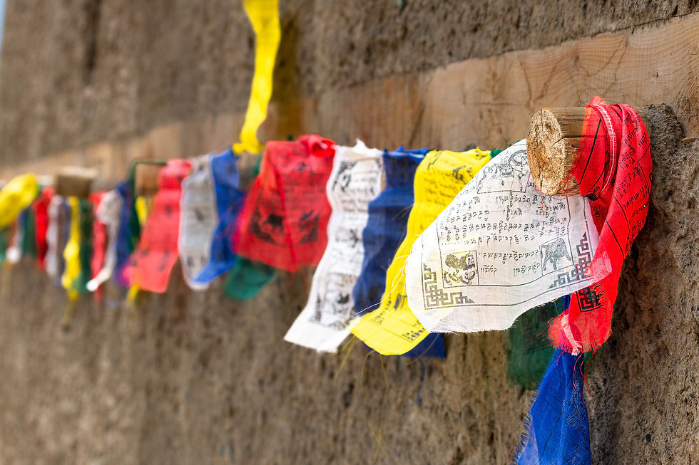Colorful prayer flags with symbols and text hang on a rough stone wall, gently blowing in the wind. The mood is calm and serene.