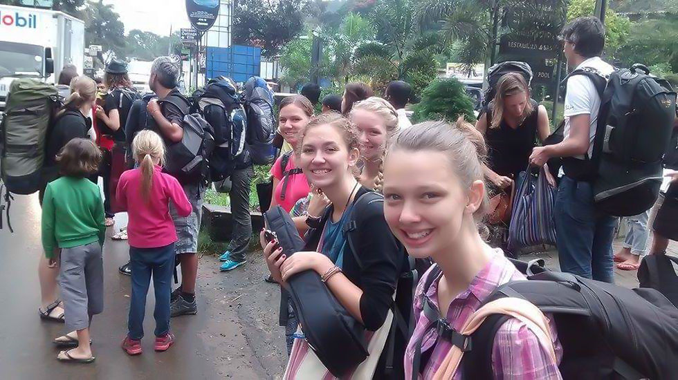 Group of backpackers smiling, standing on a rainy street with lush greenery. Visible signage and a truck in the background. Mixed emotions.