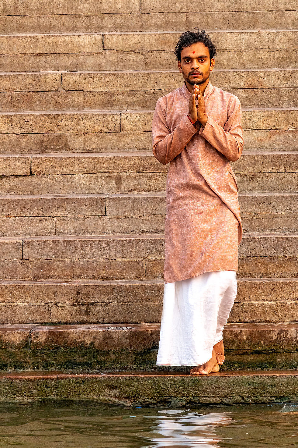 Man in traditional attire stands on stone steps near water, hands in prayer. Brown kurta, white dhoti; calm and serene atmosphere.