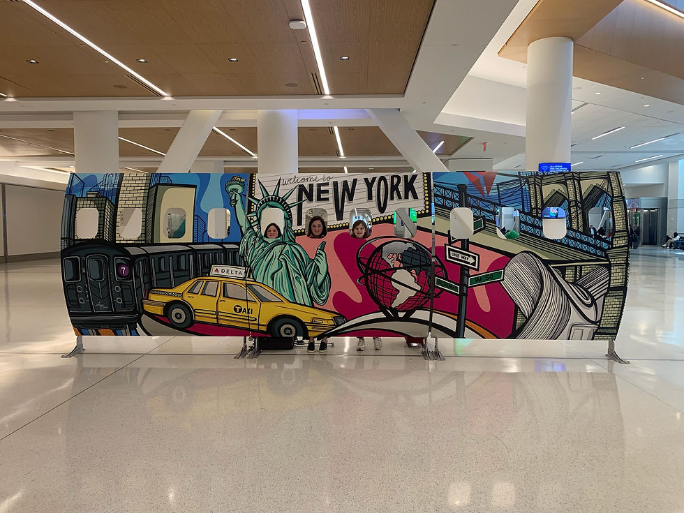 People pose behind a colorful New York-themed cutout at an airport, featuring the Statue of Liberty, subway, and a taxi. "Welcome to New York" text displayed.