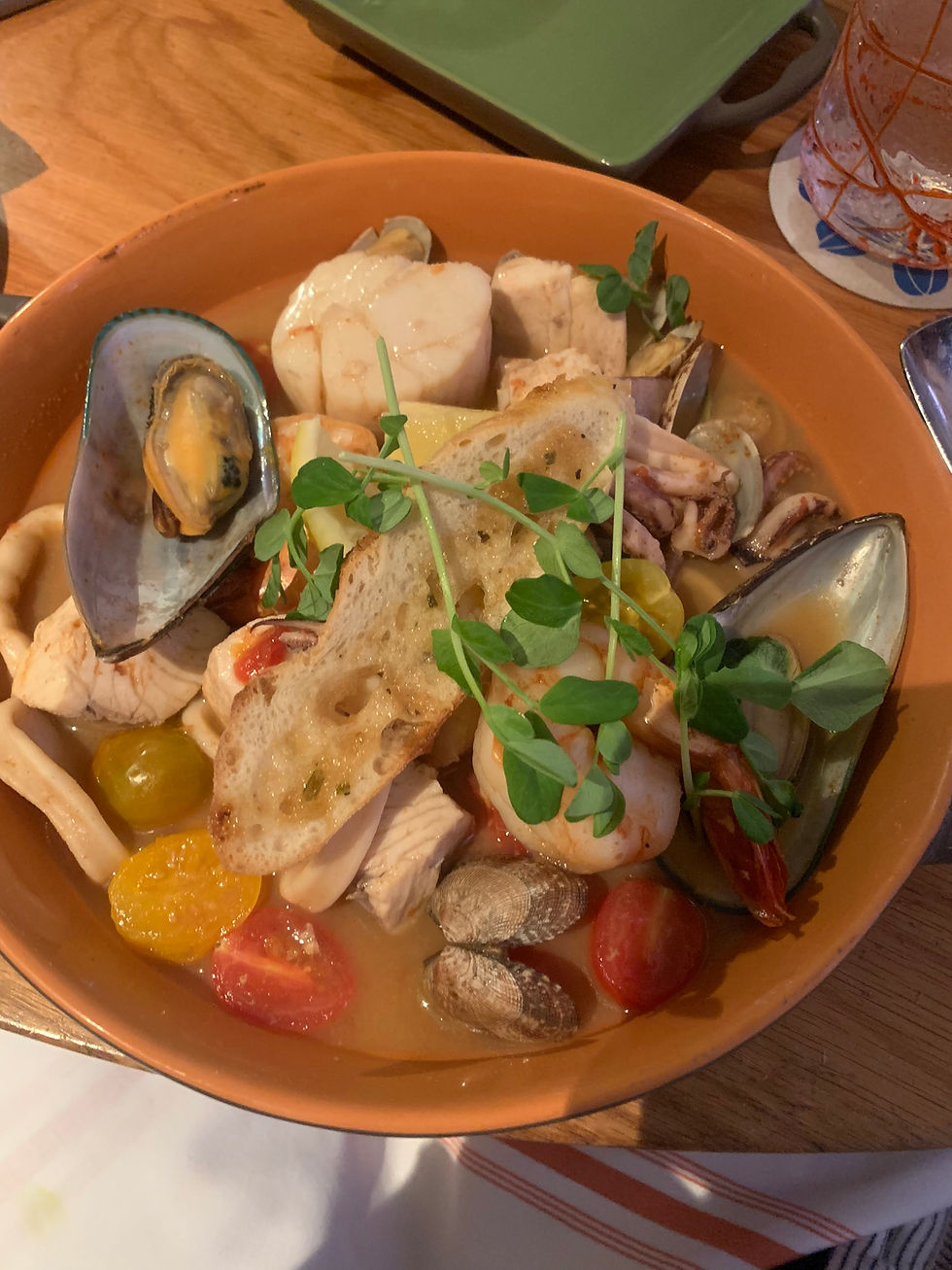 A seafood stew in a brown bowl with mussels, shrimp, tomatoes, and bread, garnished with greens, sits on a wooden table.