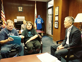 An advocate with lived experience wearing a black shirt with a chewy in his mouth and wearing glasses telling his story using an iPad held for him by his support provider a man wearing a dark blue shirt with dark hair. They are sitting across from a Legislator in the legislator's office.