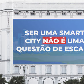 Faixa publicitária afixada na fachada de um edifício clássico com a frase "Ser uma smart city não é uma questão de escala" em letras grandes e brancas sobre um fundo azul, destacando as palavras "não é" em vermelho. A imagem comunica que a transformação em smart city depende de estratégia e não do tamanho da cidade.