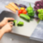 Hands slicing a cucumber on a metal cutting board. Colorful veggies like carrots, tomatoes, and lettuce on a speckled countertop. Product Photography in Delhi.