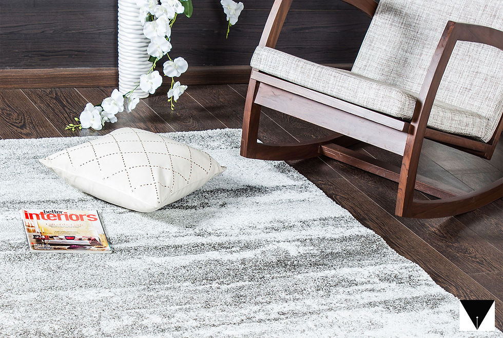 Wooden rocking chair on dark floor, with white cushion and magazine on a gray rug. White flowers in vase. Cozy and calm setting.