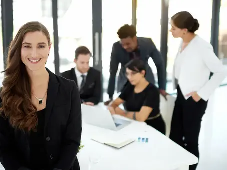 A confident, smiling female leader in a black blazer at a modern office, backed by her professional team working at a table, representing a top sales staffing agency that helps businesses optimize operations.