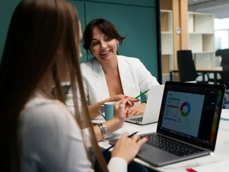 Two businesswomen collaborate in a modern office, reviewing sales performance data on a laptop, illustrating a successful partnership that scales revenue with the complete guide to sales outsourcing from Confetti Recruitment.