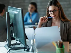 A focused female sales development representative (SDR) in glasses with a pen reviews documents at her desk, with a desktop computer and colleagues in the background, illustrating the meaning of an SDR in sales building a revenue machine.