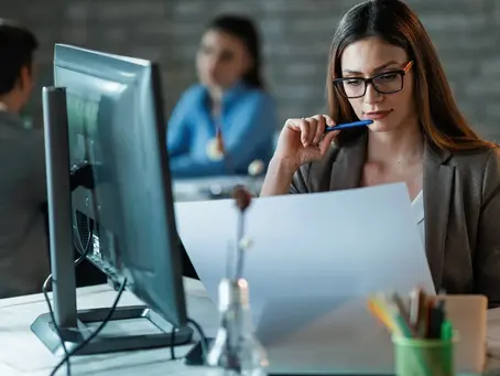 A focused female sales development representative (SDR) in glasses with a pen reviews documents at her desk, with a desktop computer and colleagues in the background, illustrating the meaning of an SDR in sales building a revenue machine.