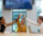 A smiling B2B sales team of three professionals gives a high five in a modern office. A monitor displaying 'IMPORT BUSINESS' and 'MARKET CENTERS' metrics confirms they are achieving goals, demonstrating the value of a top sales outsourcing company.