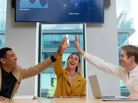 A smiling B2B sales team of three professionals gives a high five in a modern office. A monitor displaying 'IMPORT BUSINESS' and 'MARKET CENTERS' metrics confirms they are achieving goals, demonstrating the value of a top sales outsourcing company.
