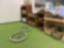 An indoor loose‑parts play area with wicker baskets, wooden shelves, natural materials, and two bicycle wheels on a green carpet