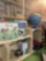 A wooden shelf with children’s books about food, a blue globe, and play kitchen items such as jars, tins, and utensils, set against a wallpaper backdrop of bookshelves