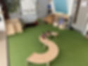 An indoor playroom with green carpet, curved wooden benches in an S‑shape, a small climbing ramp, sensory tables, and activity areas arranged around the room