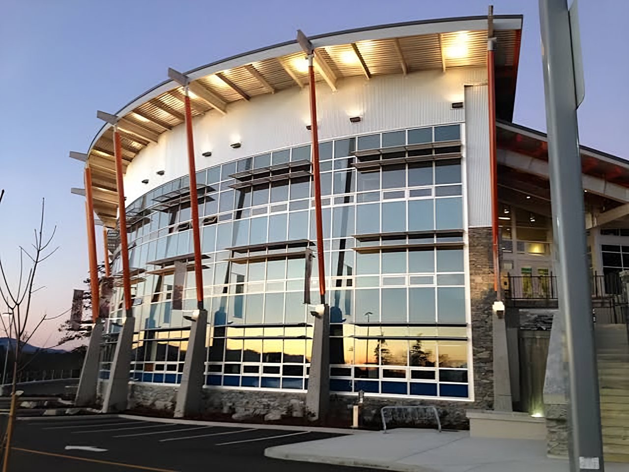 The Songhees Wellness Centre at dusk, its curved glass front lit up beneath a timber roof with red support columns