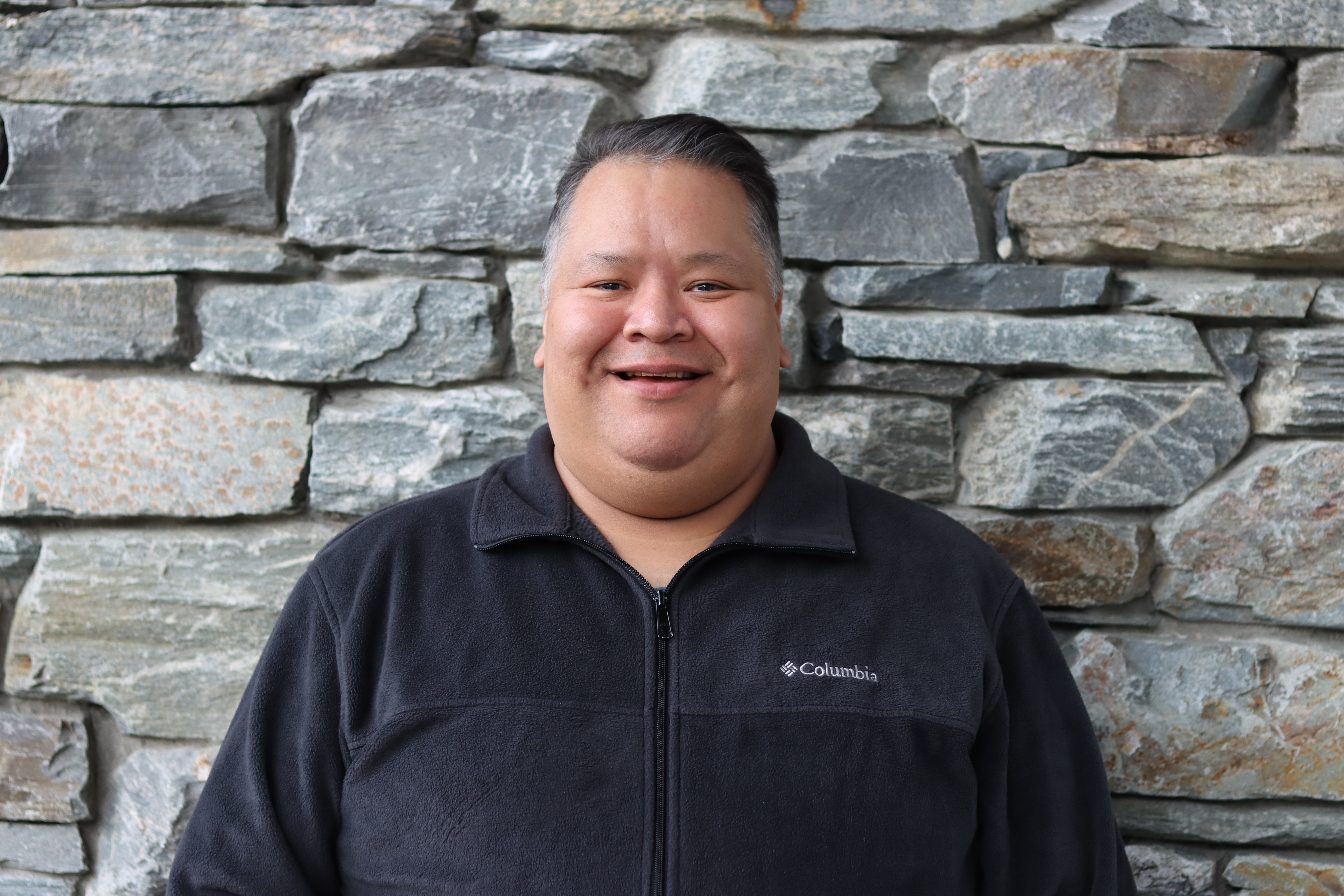 Aaron George smiling in a black fleece in front of a stone wall