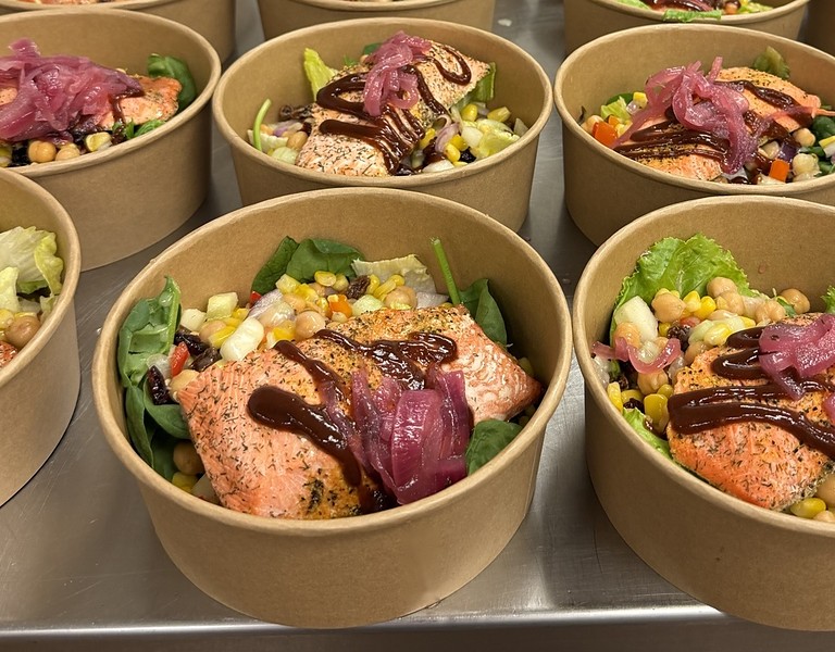 Rows of salmon bowls in kraft containers, topped with berry glaze and pickled red onion over greens, corn and chickpeas