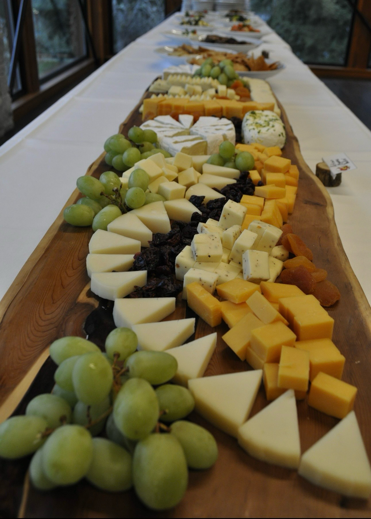 A long live-edge wooden board of sliced and cubed cheeses with green grapes, dried apricots and cranberries