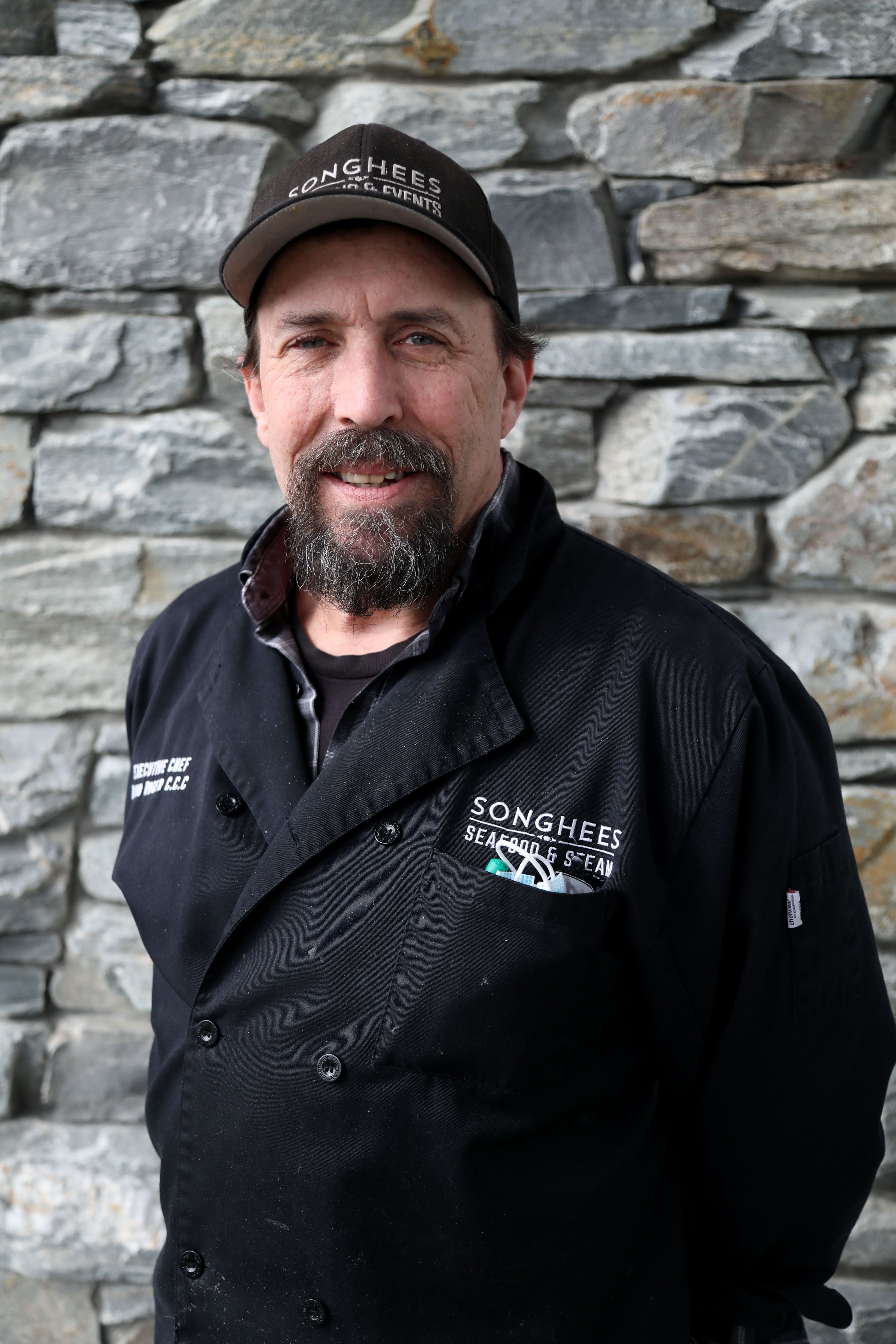 Chef David Roger in a black Songhees chef's jacket and cap, in front of a stone wall