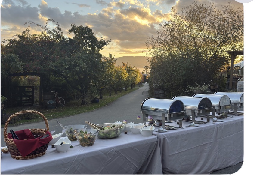 The Songhees Buffet set outdoors for a wedding along a tree-lined lane at sunset, with silver chafing dishes, salads and a basket of bannock