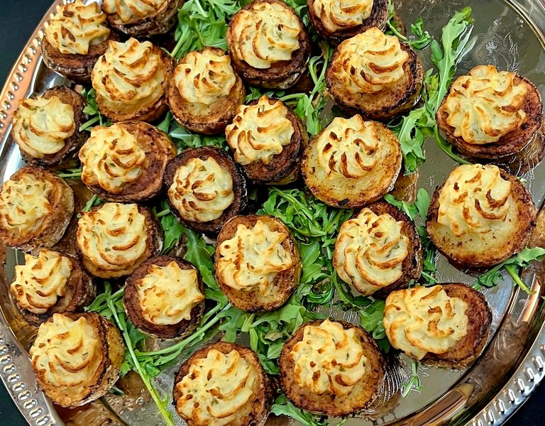 A silver platter of mini potatoes topped with golden piped filling, set on fresh arugula