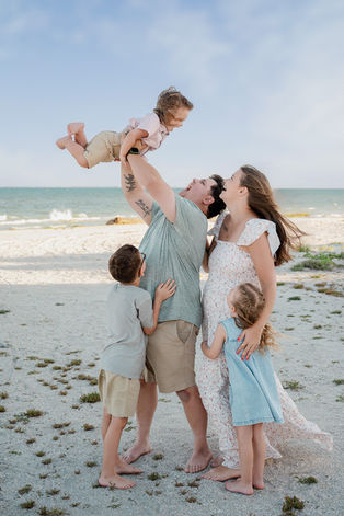 Father lifting his toddler into the air while mom and daughter smile nearby on the beach during a family photography session in Central Florida.