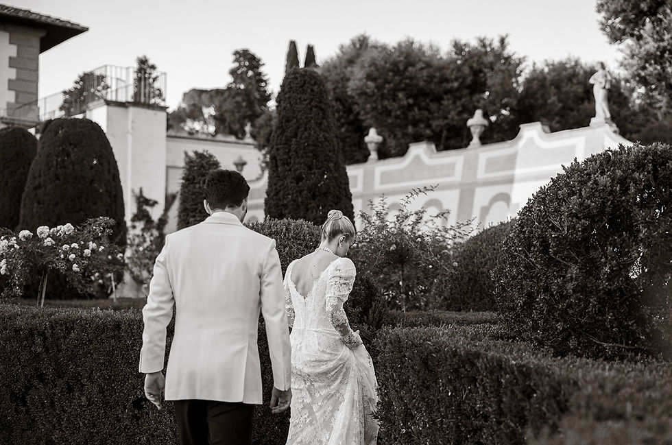 Bride in lace wedding dress and groom in white jacket walk through a manicured garden in black-and-white, calm and elegant.