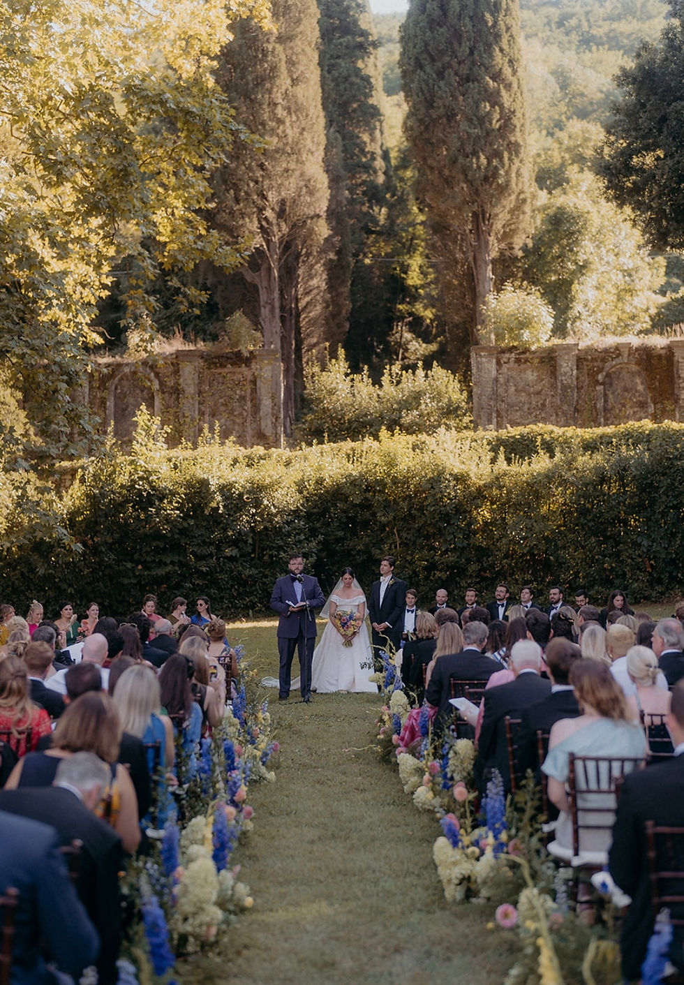 Wedding ceremony outdoors with bride, groom, and officiant. Guests seated in rows adorned with flowers. Lush greenery and trees in background.