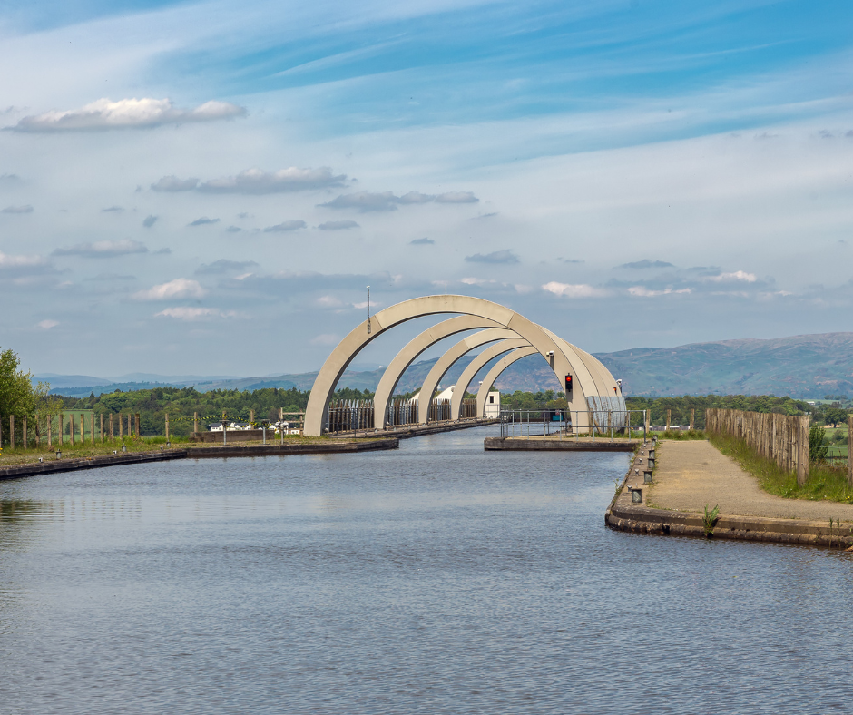 A series of large, white, arching structures rise over a calm canal under a blue sky with scattered clouds, surrounded by lush green landscape.