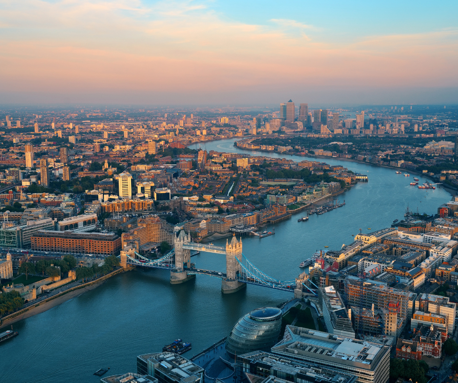 Aerial view of London at sunset, featuring the iconic Tower Bridge over the Thames River. The cityscape includes modern and historical buildings under a warm sky.