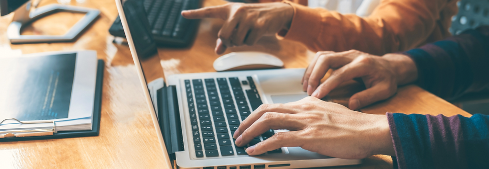 Two people work at a laptop on a wooden desk. One types while the other points at the screen. Documents and a mouse are visible. Warm tones.