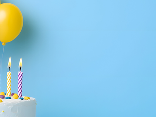 A white cake with lit striped candles, colorful candies, and pink, blue, and yellow balloons against a light blue background.
