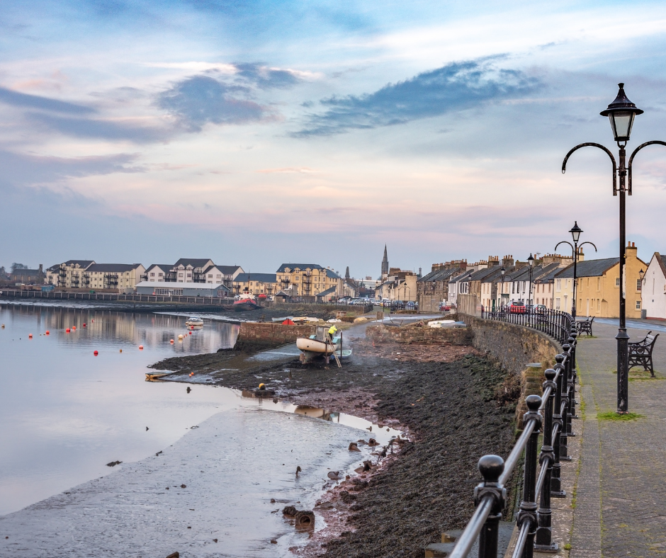 Seaside village in North Ayrshire at dawn; calm waters, docked boats, and a row of historic buildings. Street lamps illuminate a peaceful, early morning ambiance.