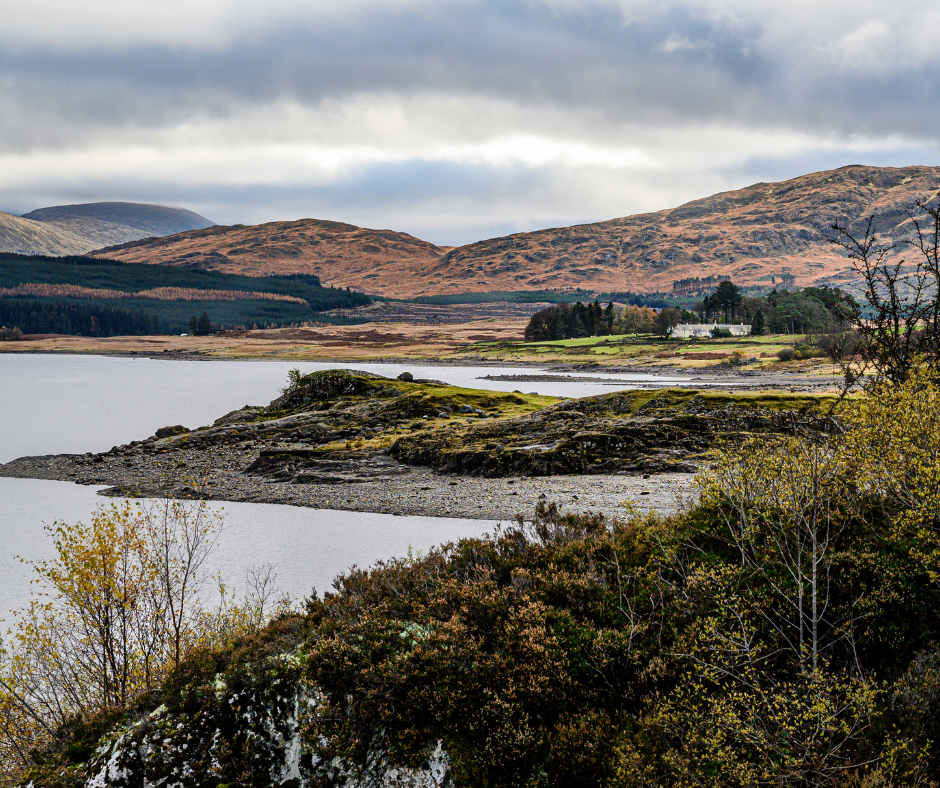 Autumn landscape with a calm lake surrounded by rocky shores and golden-brown hills under a cloudy sky. Sparse trees add to the serene atmosphere.