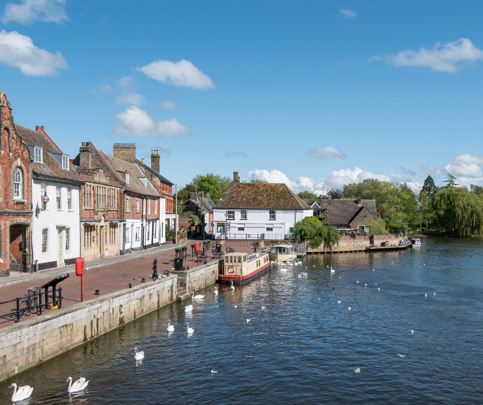 Historic riverside scene with swans, narrowboat, and charming row of brick and whitewashed buildings under a bright blue sky with fluffy clouds.