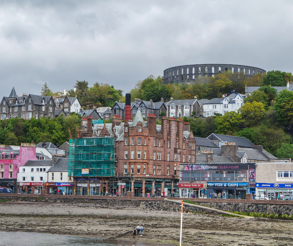 Coastal town with colorful buildings by the shore, backed by a green hillside with historic McCaig's Tower. Cloudy sky adds a calm, quaint vibe.