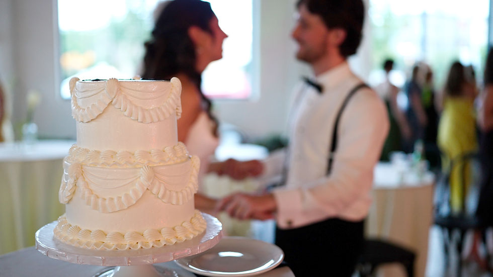 White two-tier wedding cake in foreground with blurred bride and groom dancing in a bright reception hall at the Arbor Loft at Rochester, NY.