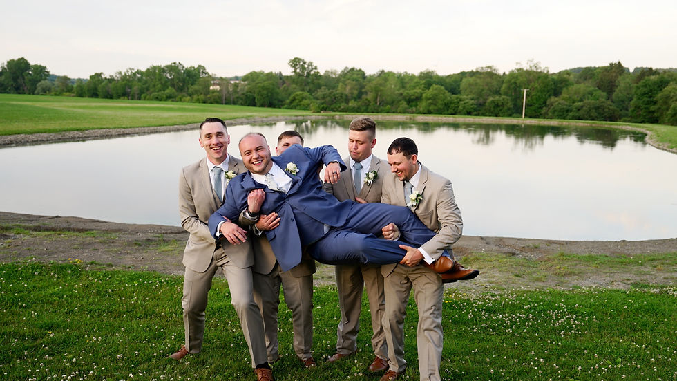 Five men in suits joyfully lift another man in a blue suit by a lake, with green grass and trees in the background. Smiling faces, festive mood at Graceful Gatherings at LedgeRock in Varysburg, NY.