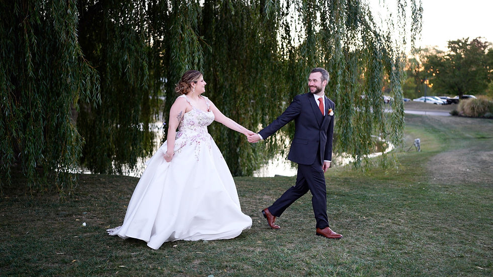 Bride in white gown and groom in dark suit joyfully holding hands, walking on grass. Weeping willow and sunset in the background at The Timberlodge at Arrowhead Golf Club in Akron, NY.